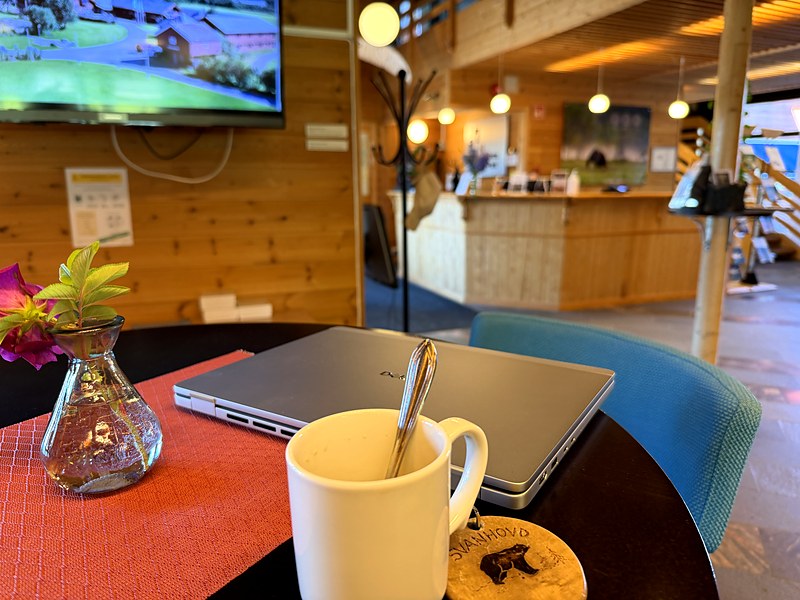 Laptop, coffee mug and the Svanhovd bear keyring on a table, with reception and the gallery level beyond